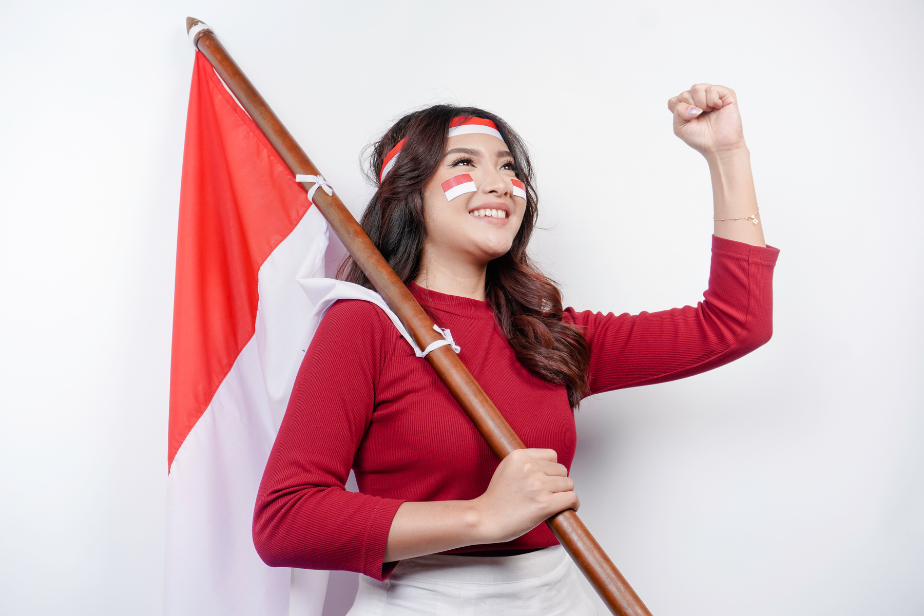 A young Asian woman with a happy successful expression wearing red top and headband while holding Indonesia's flag, isolated by white background. Indonesia's independence day concept.